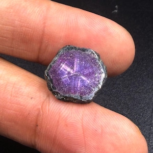 Può includere: Una fetta esagonale lucidata di una pietra preziosa viola caratterizzata da un distinto motivo a stella a sei punte che si irradia dal centro. La pietra presenta un bordo esterno scuro, ruvido e irregolare, in contrasto con l'interno liscio, traslucido e di un viola vibrante.