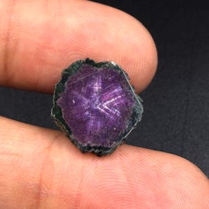 Può includere: Una fetta di pietra preziosa grezza di forma esagonale caratterizzata da un centro viola intenso con un distinto motivo a stella che si irradia dal centro. I bordi della pietra sono scuri, ruvidi e strutturati, in contrasto con la superficie viola liscia.