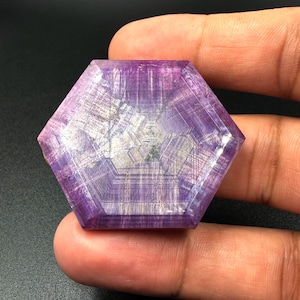 Könnte beinhalten: Ein sechseckig geschliffener violetter Edelstein mit einem ausgeprägten geometrischen Muster aus internen Linien und Facetten. Der Stein zeigt verschiedene Violett- und Magentatöne mit einer polierten Oberfläche, die das Licht auf seiner flachen Form reflektiert.