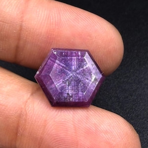 Puede incluir: Una piedra preciosa de corte hexagonal en tonos púrpura y magenta intensos, con un patrón de estrella de seis puntas visible en su superficie facetada. La piedra es translúcida y presenta inclusiones fibrosas en su interior.