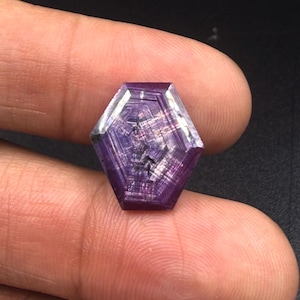 Puede incluir: Una piedra preciosa de corte hexagonal con un tono púrpura y magenta intenso, que presenta un patrón geométrico distintivo de líneas concéntricas e inclusiones internas que irradian desde el centro hacia los bordes facetados.