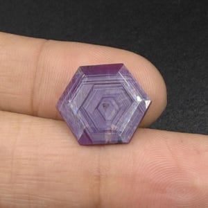 May include: A hexagonal purple gemstone featuring a distinct geometric pattern of concentric nested hexagons etched into its surface. The stone displays varying shades of violet and lavender with a polished, faceted finish and sharp edges.