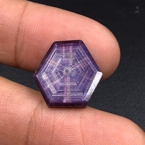 Könnte beinhalten: Ein hexagonaler violetter Edelstein mit einem ausgeprägten Trapiche-Muster, bei dem strahlenförmige Linien von einem zentralen Punkt zu den sechs Ecken verlaufen und einen geometrischen sternartigen Effekt auf der polierten Oberfläche erzeugen.