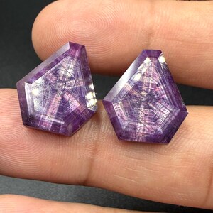 May include: Two pentagonal-shaped purple gemstones featuring a distinct star-like pattern of intersecting lines across their faceted surfaces. The stones exhibit deep violet hues with translucent qualities and a polished finish, showing internal inclusions.