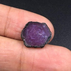 Könnte beinhalten: Ein loser, sechseckiger Edelstein in tiefem Violett und Magenta. Der Stein hat eine flache, polierte Oberfläche mit sichtbaren inneren Kristallstrukturen und einen dunklen, rauen, natürlichen Rand um den gesamten Umfang.