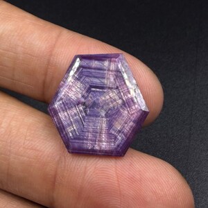 Può includere: Una gemma esagonale viola caratterizzata da un distinto motivo geometrico di linee concentriche che si irradiano dal centro. La pietra mostra una qualità traslucida con striature interne visibili e una consistenza superficiale lucida.