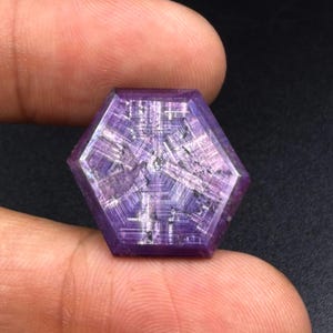 Può includere: Una gemma esagonale viola caratterizzata da un distinto motivo a stella con linee bianche incrociate che si irradiano dal centro. La pietra presenta una superficie lucida con inclusioni interne visibili e una tonalità viola intenso su tutte le faccette.