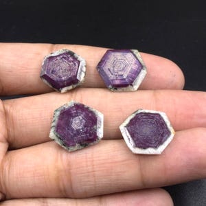 Puede incluir: Cuatro piedras preciosas de rubí de forma hexagonal con patrones geométricos distintos en tonos magenta y púrpura profundos. Cada piedra muestra una estructura cristalina natural con inclusiones minerales blancas y grises en los bordes exteriores.