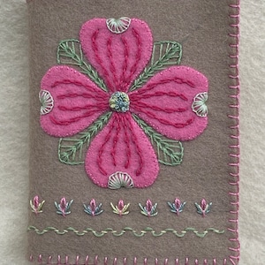 May include: A brown felt needle case with a pink flower embroidered on the front. The flower has a green stem and leaves. There are smaller pink flowers embroidered along the bottom edge.