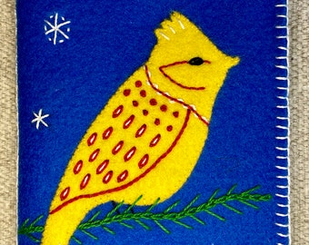 May include: A yellow felt bird with red and white accents sits on a green felt branch against a blue felt background with white snowflakes.
