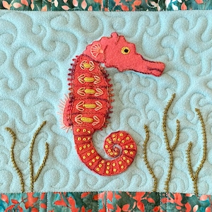 May include: A pink and gold embroidered seahorse with a curled tail, surrounded by gold embroidered seaweed on a light blue background.