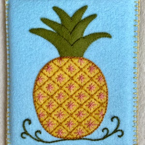 May include: A blue felt pouch with a yellow and pink embroidered pineapple design. The pineapple has green leaves and a brown stem.
