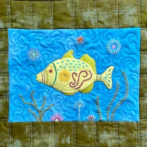 May include: A yellow and blue embroidered fish with a swirl design on a blue background with green seaweed and white flowers. The fish has a pink eye and a yellow and green tail.