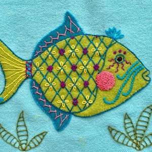 May include: A hand-stitched embroidered fish with a yellow body, blue and pink details, and a turquoise background. The fish is surrounded by green embroidered plants.