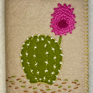 May include: A felt craft depicting a green cactus with white flowers and a pink flower on top. The cactus is surrounded by brown and green stitches on a beige background.