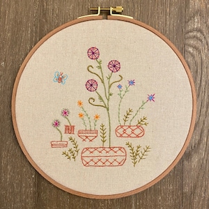 Puede incluir: Arte de bordado en aro con un diseño floral con flores rosas, azules y verdes en macetas de terracota. El diseño está bordado sobre un fondo de tela beige.