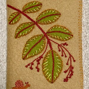 May include: A beige felt fabric pouch with a stitched border. The pouch features an embroidered design of a green leaf branch with red berries and a small snail in the bottom corner.