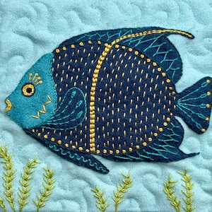 May include: A blue and gold embroidered fish with a detailed design, on a light blue quilted fabric background.