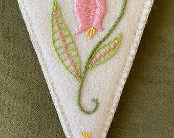 May include: A white felt triangle with a pink flower and green leaves embroidered on it. The flower has a yellow center and the leaves have pink dots. There are also yellow star-shaped flowers embroidered on the triangle.