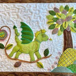 Könnte beinhalten: Ein handgefertigtes Textilkunstwerk mit einem grünen Drachen mit gelben Akzenten, der auf einem braunen Ast sitzt. Rechts befindet sich ein Baum mit grünen, gelben und grauen Blättern. Der Hintergrund ist weiß mit einem gesteppten Muster.