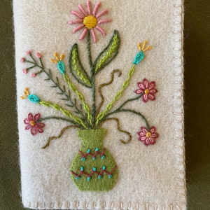 Puede incluir: Una aguja de fieltro blanco con un jarrón verde lleno de flores bordadas en rosa, amarillo y verde.
