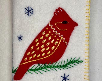 May include: A white felt cover with a red cardinal bird design. The bird is perched on a green branch, with gold and white embroidered details. The edges are stitched with gold thread, and blue snowflakes are scattered around the bird.