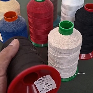 Op de afbeelding: Een verzameling industriële naaigarenspoelen in diverse kleuren, waaronder rood, wit, zwart, blauw, beige en bruin. De spoelen hebben kunststof kernen en voetjes. Eén spoel die op de voorgrond wordt vastgehouden, toont een label met POLICON 10/3.