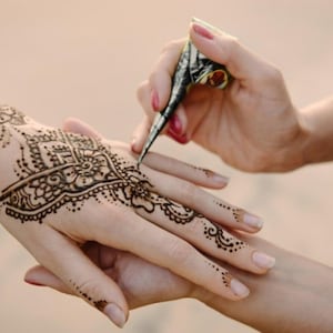Può includere: Una mano viene decorata con intricati disegni all'henné utilizzando un applicatore a cono. Il motivo all'henné è marrone scuro e presenta elementi floreali e geometrici. L'incarnato è chiaro e lo sfondo è di un colore neutro.