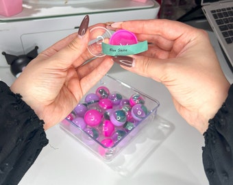 May include: A clear plastic container filled with pink, purple, and clear marbles. A small, clear plastic lid holds a pink object with a green label that reads "Blue Shine".