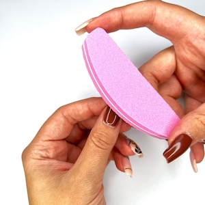 May include: A pink nail file with white edges is held between two hands. The file is being used on a nail with a brown manicure. The background is white.