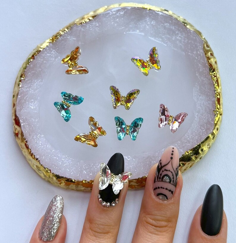 May include: A collection of iridescent butterfly nail art decorations in various colors, including gold, teal, and pink, are displayed on a white and gold-edged surface. The image also shows a hand with several manicured nails, some with glitter and decorative elements.