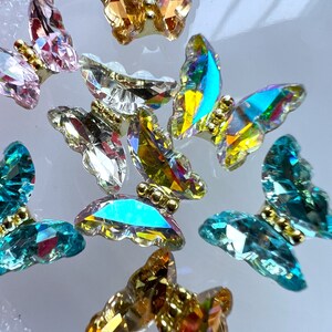 May include: Close-up of several iridescent butterfly-shaped crystal embellishments. The crystals are in various colors, including pink, gold, teal, and clear. Each butterfly has a gold-colored body.