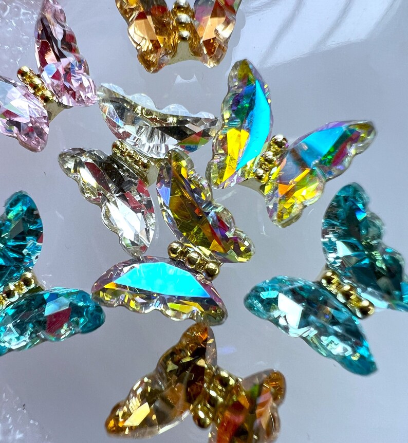 May include: Close-up of several iridescent butterfly-shaped crystal embellishments. The crystals are in various colors, including pink, gold, teal, and clear. Each butterfly has a gold-colored body.