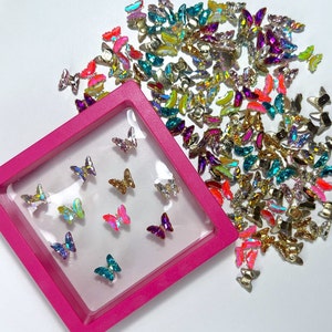 May include: A collection of colorful butterfly-shaped nail art decorations. The butterflies are in various colors, including pink, gold, teal, and purple, and are displayed in a pink square container and scattered on a white surface.