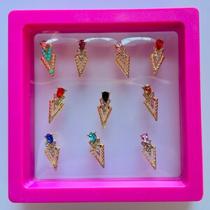 May include: A collection of gold-colored arrow-shaped nail charms with various colored gemstones, including turquoise, red, and pink. The charms are displayed in a pink rectangular box, creating a vibrant and eye-catching presentation. These nail art accessories add a touch of glamour.