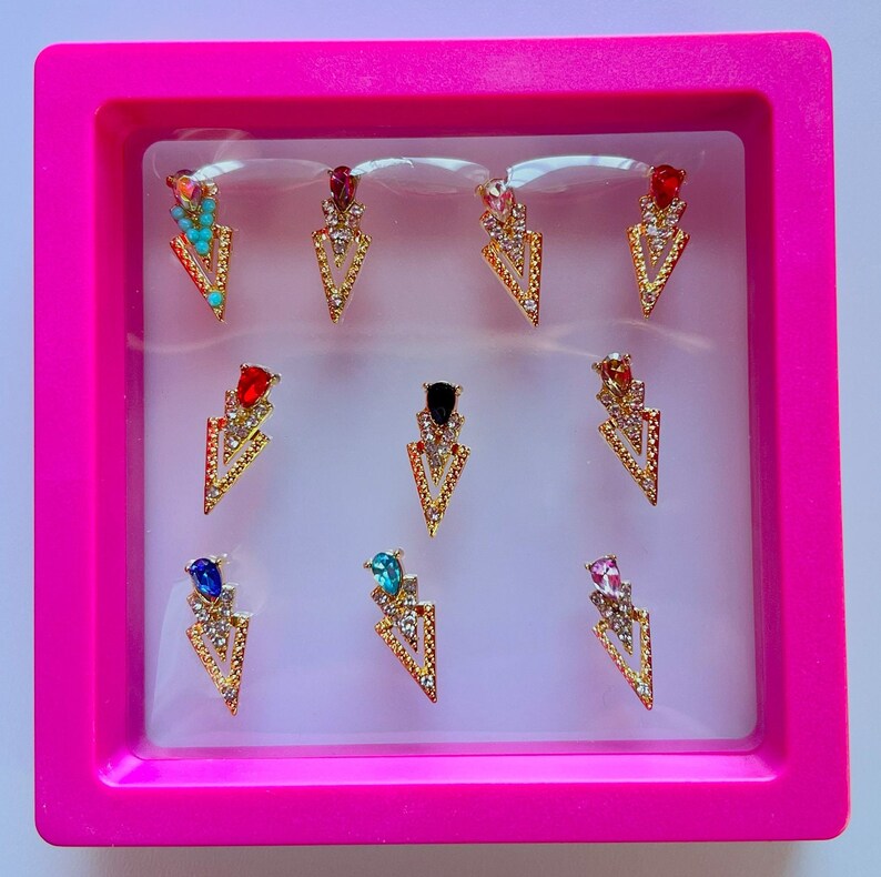 May include: A collection of gold-colored arrow-shaped nail charms with various colored gemstones, including turquoise, red, and pink. The charms are displayed in a pink rectangular box, creating a vibrant and eye-catching presentation. These nail art accessories add a touch of glamour.