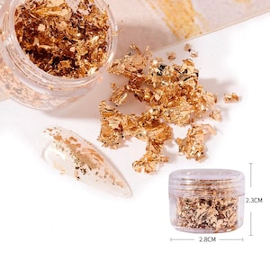 May include: Gold leaf flakes in a clear plastic jar, with a nail art sample. The jar is 2.8 cm wide and 2.3 cm tall. Scattered gold flakes are also visible. Ideal for nail art and craft projects.