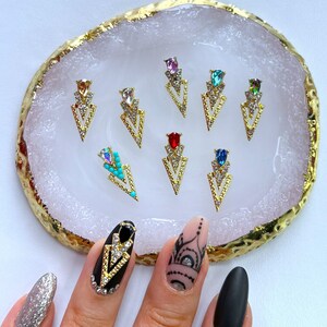 May include: A collection of gold-colored nail art charms in a variety of jewel tones, including red, blue, and turquoise. The charms are arranged on a white and gold decorative tray. The image also shows a hand with decorated nails.