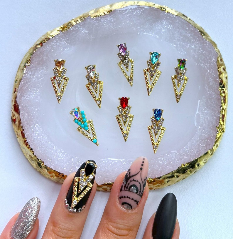 May include: A collection of gold-colored nail art charms in a variety of jewel tones, including red, blue, and turquoise. The charms are arranged on a white and gold decorative tray. The image also shows a hand with decorated nails.