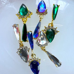 May include: A collection of ornate, dangling earrings with a variety of jewel-toned gemstones. The earrings feature emerald green, sapphire blue, and clear crystals set in gold-colored metal. The gemstones are teardrop and oval shaped.