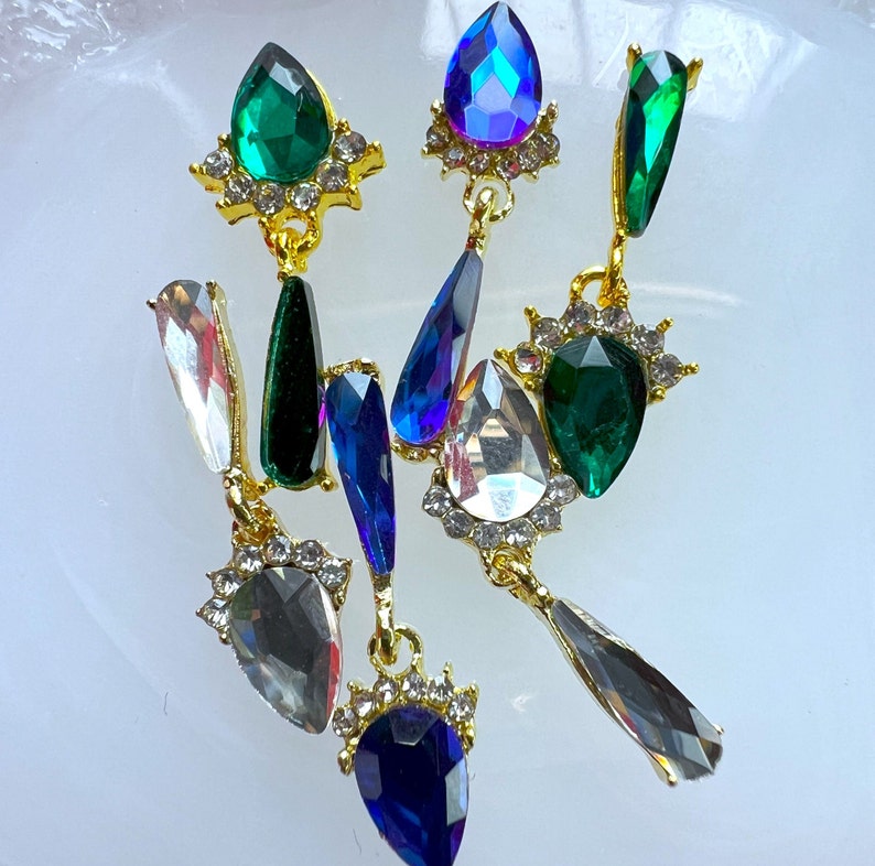 May include: A collection of ornate, dangling earrings with a variety of jewel-toned gemstones. The earrings feature emerald green, sapphire blue, and clear crystals set in gold-colored metal. The gemstones are teardrop and oval shaped.