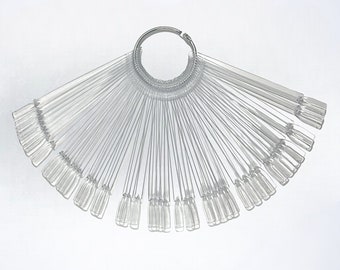 May include: A clear plastic nail polish color swatch fan with a silver metal ring. Each swatch is a rectangular shape with a rounded tip. The fan is spread out, displaying multiple color options for nail polish.