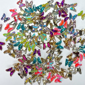 May include: A collection of iridescent butterfly-shaped embellishments in various colors, including pink, teal, gold, and purple. These decorative pieces have a metallic base and are scattered across a white surface, ideal for crafts and embellishments.