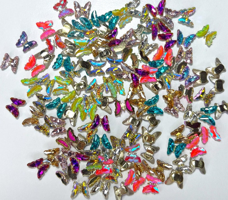 May include: A collection of iridescent butterfly-shaped embellishments in various colors, including pink, teal, gold, and purple. These decorative pieces have a metallic base and are scattered across a white surface, ideal for crafts and embellishments.