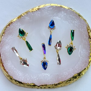 May include: An assortment of gemstone earrings in various colors, including green, blue, and clear, are displayed on a pink and gold-edged tray. Each earring features a teardrop-shaped gem and a decorative gold setting.