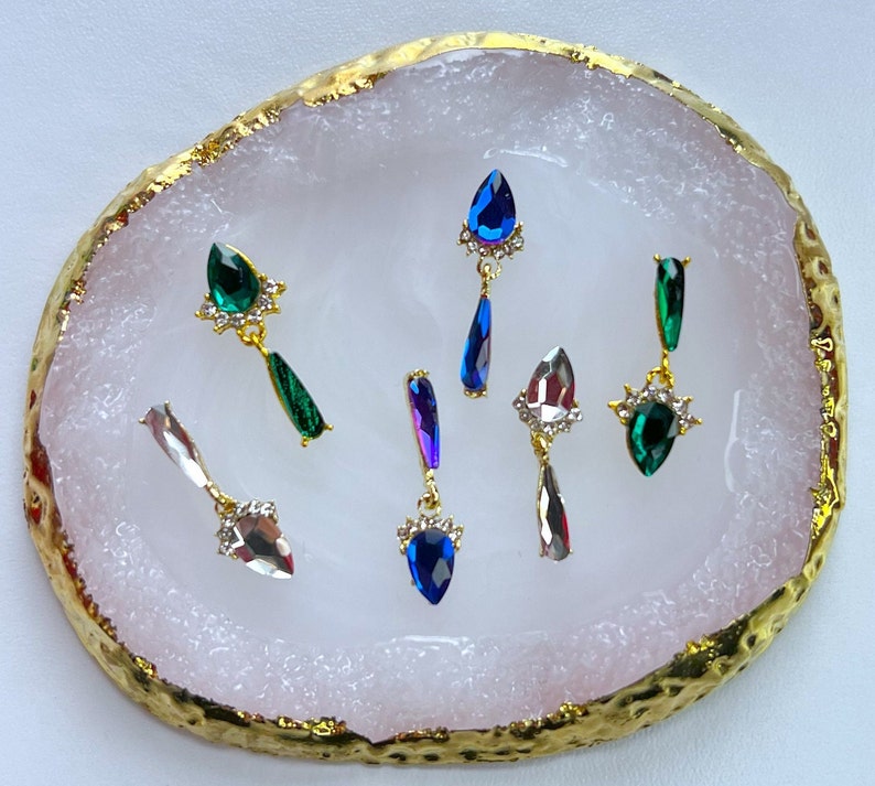 May include: An assortment of gemstone earrings in various colors, including green, blue, and clear, are displayed on a pink and gold-edged tray. Each earring features a teardrop-shaped gem and a decorative gold setting.