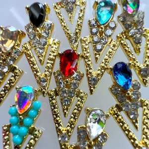 May include: Close-up of several gold-colored, arrow-shaped jewelry pieces. Each piece features a colorful gemstone at the top, including black, red, blue, and iridescent options, surrounded by small, clear crystals.