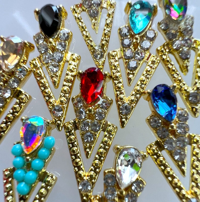 May include: Close-up of several gold-colored, arrow-shaped jewelry pieces. Each piece features a colorful gemstone at the top, including black, red, blue, and iridescent options, surrounded by small, clear crystals.