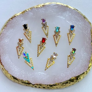 May include: A collection of gold-colored arrow-shaped nail art charms. Each charm features a central gemstone in various colors, including black, red, and teal, surrounded by clear crystals. The charms are arranged on a white and gold-edged surface.