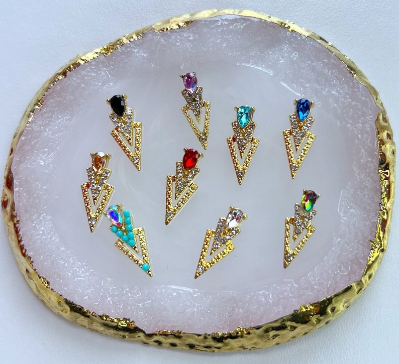 May include: A collection of gold-colored arrow-shaped nail art charms. Each charm features a central gemstone in various colors, including black, red, and teal, surrounded by clear crystals. The charms are arranged on a white and gold-edged surface.
